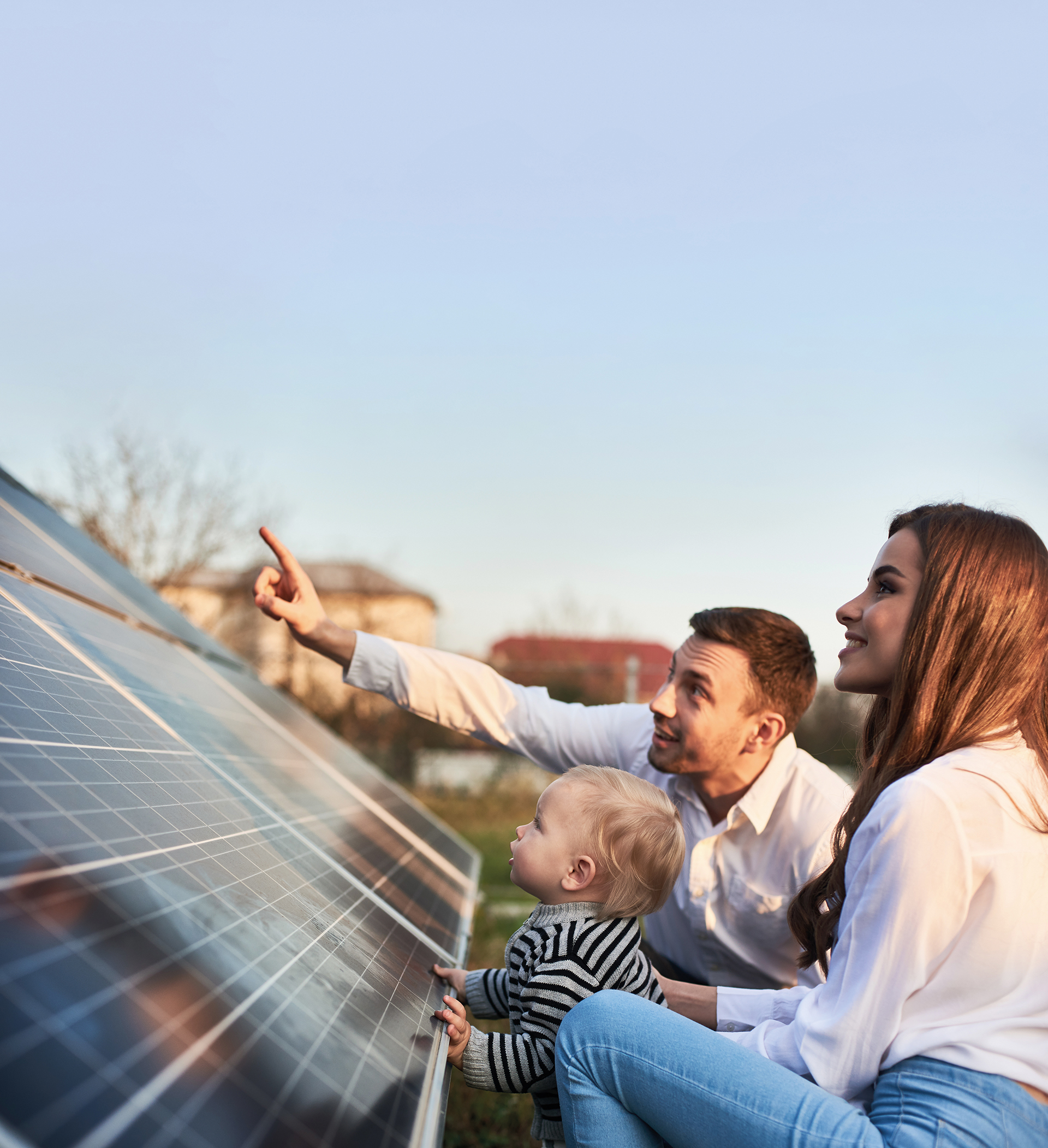 Man shows his family the solar panels on the plot near the house during a warm day. Young woman with a kid and a man in the sun rays look at the solar panels.