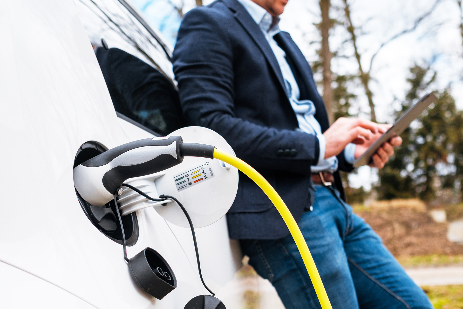Business man standing near charging electric car or EV car and using tablet in the street