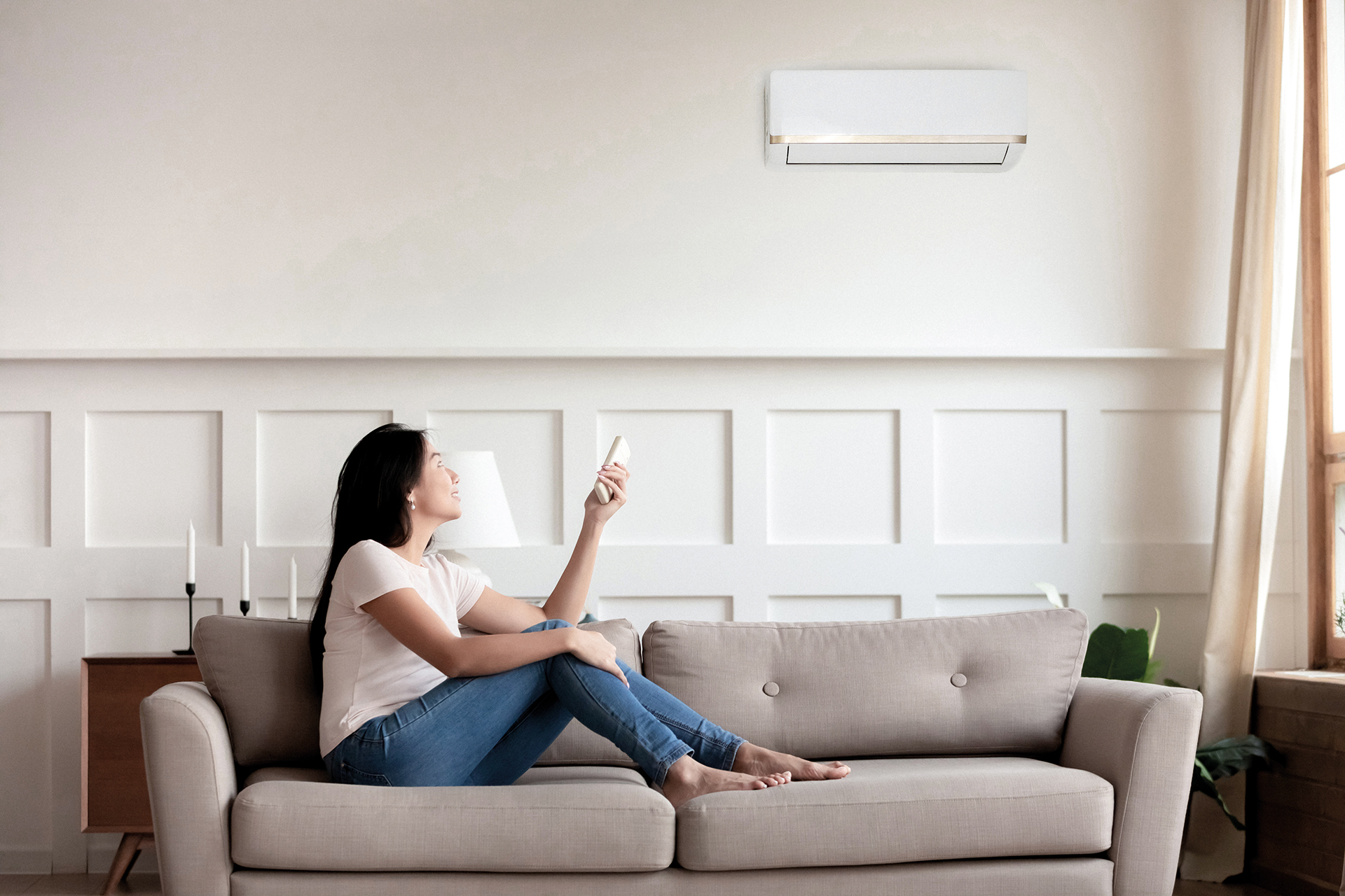 Vietnamese young woman relaxing on comfy couch in contemporary living room with air conditioner, holding remote control turning on or off cooler system, setting comfortable temperature at modern home